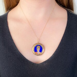 Pu&ograve; includere: Una collana con catena d'oro con un ciondolo rotondo a forma di medaglione. Il medaglione presenta un ritratto dipinto di una donna con una corona di fiori su sfondo blu.