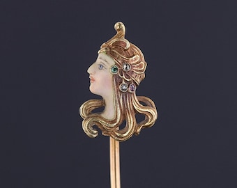 May include: A gold stick pin with a detailed enamel portrait of a woman with a crown and colorful gemstones. The woman's hair is styled in a wavy design.