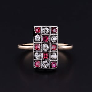 May include: A rectangular gold ring featuring a grid of alternating red and clear gemstones. The gemstones are square-cut and set in a silver-colored metal. The ring has a simple, elegant design.