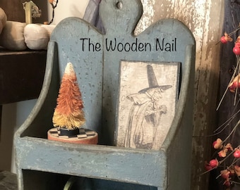 Può includere: Uno scaffale in legno blu a due ripiani. Il ripiano superiore presenta un piccolo albero di mais dolce arancione e bianco e una stampa vintage di Halloween. Il ripiano inferiore contiene tre piccole zucche. Lo scaffale ha un cartello con la scritta "The Wooden Nail".
