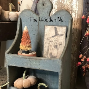 May include: A blue wooden shelf with two shelves. The top shelf has a small orange and white candy corn tree and a vintage Halloween print. The bottom shelf has three small pumpkins. The shelf has a sign that says "The Wooden Nail".