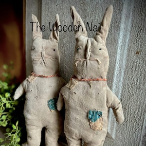 Peut inclure: Deux lapins en tissu faits à la main avec de longues oreilles, un corps brun et des liens en ficelle rouge. Les lapins ont des patchs en tissu bleu et blanc sur leur corps. Les lapins sont debout sur une surface brune devant une porte en bois blanc avec le texte "The Wooden Nail" écrit dessus.