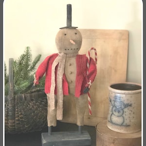 May include: A handmade primitive snowman doll with a red jacket, a tan scarf, and a black top hat. The snowman is holding a candy cane and standing on a wooden base.