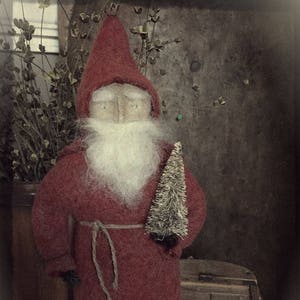 May include: A handmade felt Santa Claus doll wearing a red robe with a white beard and holding a small, sparkly Christmas tree. The doll is standing on a wooden surface with the text "The Wooden Nail" visible in the bottom right corner.