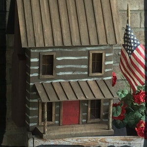 May include: A miniature wooden log cabin with a red door, white painted logs, and a metal roof. The cabin is decorated with an American flag and red flowers.