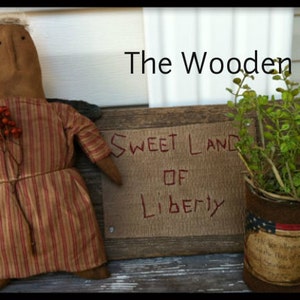 May include: A handmade rag doll with a striped dress and a sprig of red berries. The doll is standing in front of a wooden sign that reads "Sweet Land of Liberty" and "The Wooden Nail".