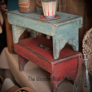Może przedstawiać: Dwa drewniane stołki ułożone jeden na drugim. Górny stołek jest pomalowany na jasnoniebieski kolor z efektem postarzenia. Dolny stołek jest pomalowany na czerwono z efektem postarzenia. Mały papierowy kapelusz w czerwono-biało-niebieskie paski leży na górnym stołku. Tekst "The Wooden Nail" jest widoczny w prawym dolnym rogu obrazu.