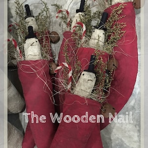 May include: Handmade Christmas decorations featuring snowmen with black top hats and carrot noses, nestled in red fabric stockings. Accented with sprigs of greenery, candy canes, and small bells. The text "The Wooden Nail" is visible.