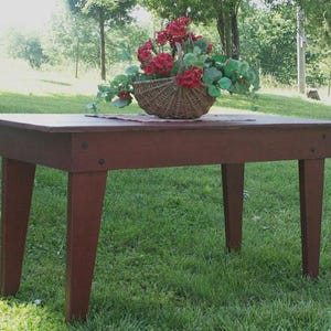Könnte beinhalten: Ein roter Holztisch mit vier Beinen und einer rechteckigen Tischplatte. Der Tisch steht auf einem Rasen mit einem Blumenarrangement in einem Weidenkorb auf der Tischplatte.
