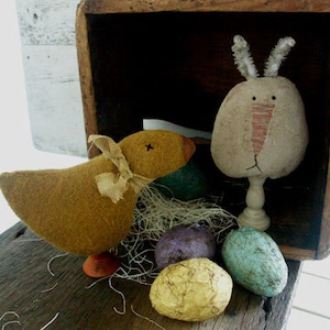 May include: A brown felt chick with a yellow beak and brown felt wings sits in a wooden crate with a felt bunny and Easter eggs. The bunny is white with a pink nose and long ears. The eggs are blue, purple, and gold.