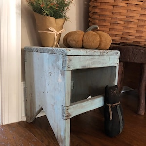 Puede incluir: Un taburete de madera azul claro con un acabado desgastado. El taburete tiene un diseño sencillo con un solo estante y una base ancha. Una muñeca de gato negro está sentada en el suelo junto al taburete.