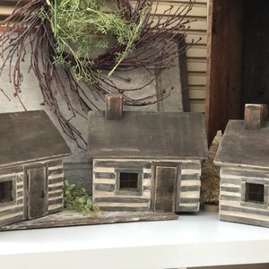 Pode incluir: Três casas de madeira em miniatura no estilo de cabana de troncos com detalhes pintados de branco. As cabanas têm um telhado de madeira marrom, uma porta e uma janela. As cabanas estão dispostas em fila sobre uma superfície branca.