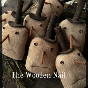 May include: A group of handmade felt snowman ornaments with black top hats and brown yarn noses. The ornaments are arranged on a wooden surface with green pine branches. The text "The Wooden Nail" is visible in the bottom center of the image.