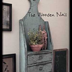 Op de afbeelding: Een lichtblauw houten wandrek met twee laden en een bovenste plank. Het rek heeft een verweerde afwerking en is versierd met een kleine plant in een pot en een houten konijnenbeeldje. De tekst "The Wooden Nail" staat op het rek.
