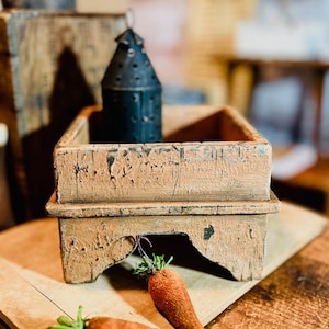 May include: A rustic wooden box with a distressed finish, featuring a raised base and a square top. The box is painted in a light brown color with hints of green and black. The box is sitting on a wooden surface with two carrots in the foreground.
