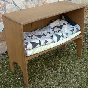 May include: A wooden shelf with a quilt folded on top. The shelf is painted a light brown color and has a simple design. The shelf is sitting on a bed of green grass.