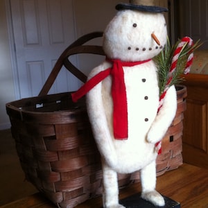 Könnte beinhalten: Ein weißer Filz-Schneemann mit einem schwarzen Zylinder, einem roten Schal und einem Zuckerstangen in der Hand. Der Schneemann steht auf einem Holzsockel mit schwarzer Oberseite.