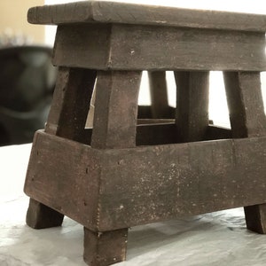 May include: A dark brown wooden stool with a square top and four legs. The stool has a simple, rustic design.