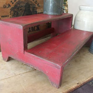 May include: A two-step red wooden stool with a distressed finish. The stool has a simple design with rounded edges and a slightly raised lip on the top step.