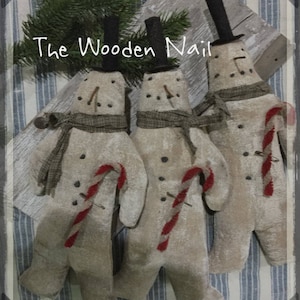 May include: Three handmade fabric snowmen with black top hats, brown scarves, and red and white candy canes. The snowmen are standing on a striped fabric background. The text "The Wooden Nail" is visible at the top of the image.