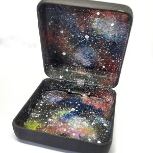 May include: A small black box with a hinged lid. The inside of the box is painted to look like a galaxy with stars and nebulae.