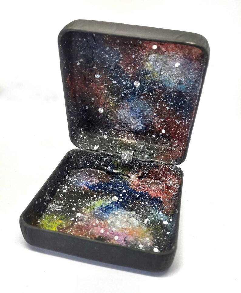 May include: A small black box with a hinged lid. The inside of the box is painted to look like a galaxy with stars and nebulae.