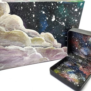 May include: A small black box with a galaxy design on the inside and a larger canvas painting with a galaxy design and white clouds.