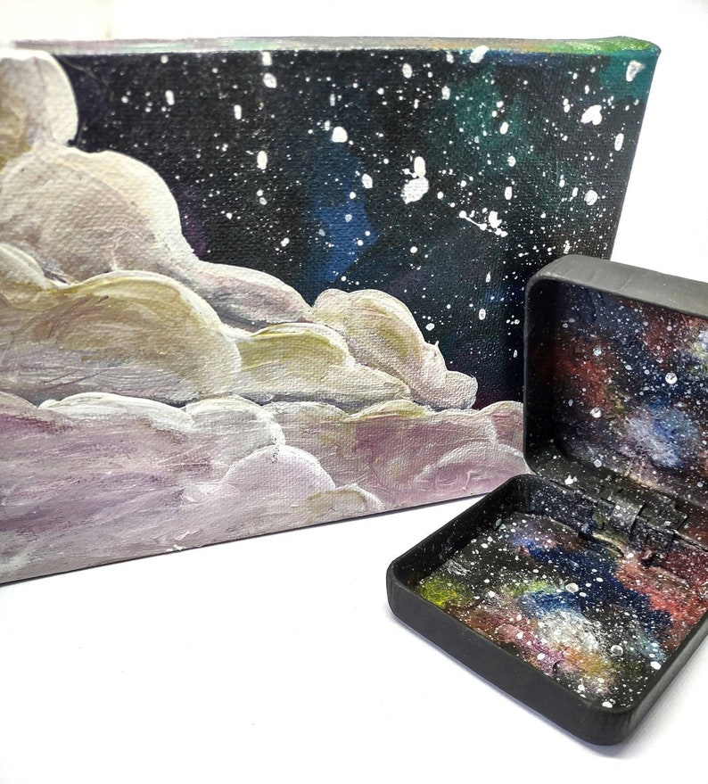 May include: A small black box with a galaxy design on the inside and a larger canvas painting with a galaxy design and white clouds.