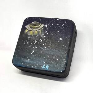 May include: A small wooden box with a black background and a painted image of a UFO flying over a starry night sky. The box is signed "LB" in the bottom right corner.