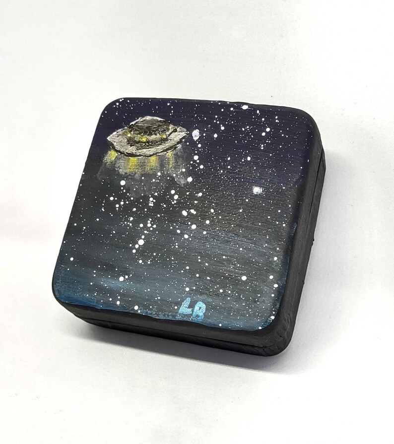 May include: A small wooden box with a black background and a painted image of a UFO flying over a starry night sky. The box is signed "LB" in the bottom right corner.