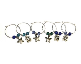 May include: Set of six silver wine charms with blue and green beads. Each charm features a different beach-themed pendant: starfish and flip-flops. These charms are perfect for a beach-themed party or a nautical event.