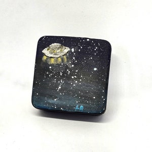 May include: A small, square, black box with a painted image of a UFO flying over a starry night sky. The UFO is white with yellow lights. The box has a blue inscription on the bottom.
