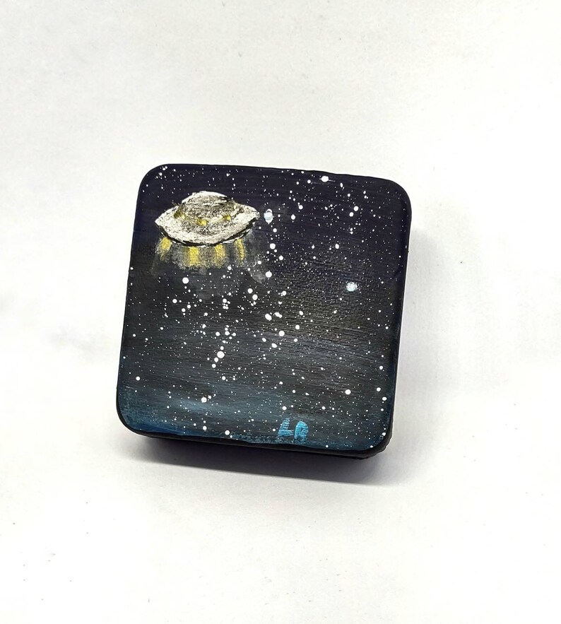 May include: A small, square, black box with a painted image of a UFO flying over a starry night sky. The UFO is white with yellow lights. The box has a blue inscription on the bottom.