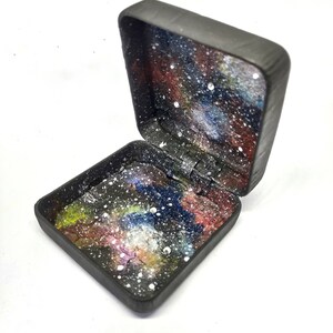 May include: A small black box with a hinged lid. The inside of the box is painted with a galaxy design featuring a dark blue, purple, and red nebula with white stars.
