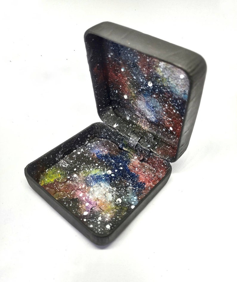 May include: A small black box with a hinged lid. The inside of the box is painted with a galaxy design featuring a dark blue, purple, and red nebula with white stars.
