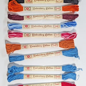 Könnte beinhalten: Eine Sammlung von Stickgarn aus Baumwolle in verschiedenen Farben, darunter Schwarz, Orange, Burgunder, Blau, Rosa und Rot. Jede Strähne ist mit dem Markennamen und der Farbnummer versehen. Ideal für Stickprojekte.