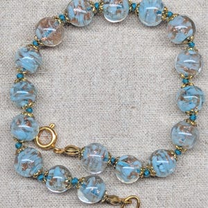 May include: A beaded bracelet featuring clear, round beads with a light blue and copper-colored swirl design. The beads are strung together with teal-colored thread and gold-toned metal accents. The bracelet is approximately 7 inches in length.