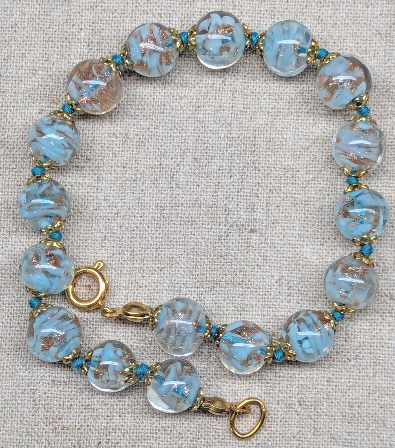 May include: A beaded bracelet featuring clear, round beads with a light blue and copper-colored swirl design. The beads are strung together with teal-colored thread and gold-toned metal accents. The bracelet is approximately 7 inches in length.