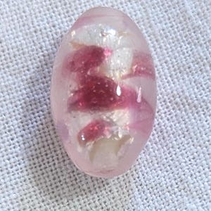 May include: Oval-shaped, translucent pink bead with embedded white and deep pink floral-like inclusions. The bead is smooth and polished, reflecting light. The design suggests a handcrafted item, possibly for jewelry or decorative purposes.