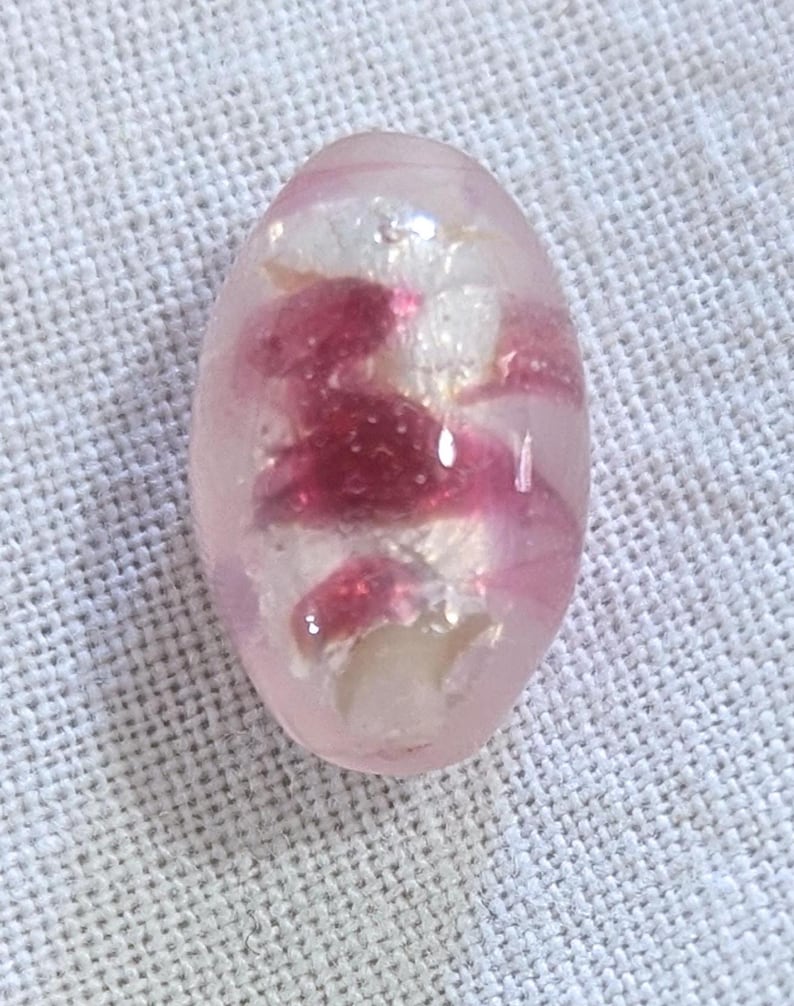 May include: Oval-shaped, translucent pink bead with embedded white and deep pink floral-like inclusions. The bead is smooth and polished, reflecting light. The design suggests a handcrafted item, possibly for jewelry or decorative purposes.