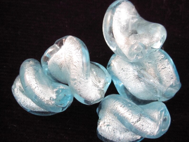 May include: Several light blue glass beads with a silver foil effect are arranged against a black background. The beads have a twisted, organic shape, and the silver foil adds a reflective quality to the translucent glass.