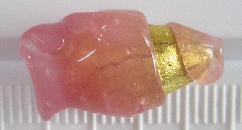 May include: A close-up of a small, abstract sculpture. The sculpture features a pink, translucent, organic shape connected to a gold-colored, cylindrical element. The piece is set against a white background with a ruler for scale.