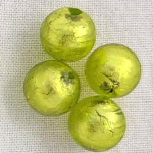 May include: Four round, yellow-green decorative beads with a metallic finish. The beads have a slightly irregular surface and appear to be made of glass or a similar material. They are arranged on a white, textured background.