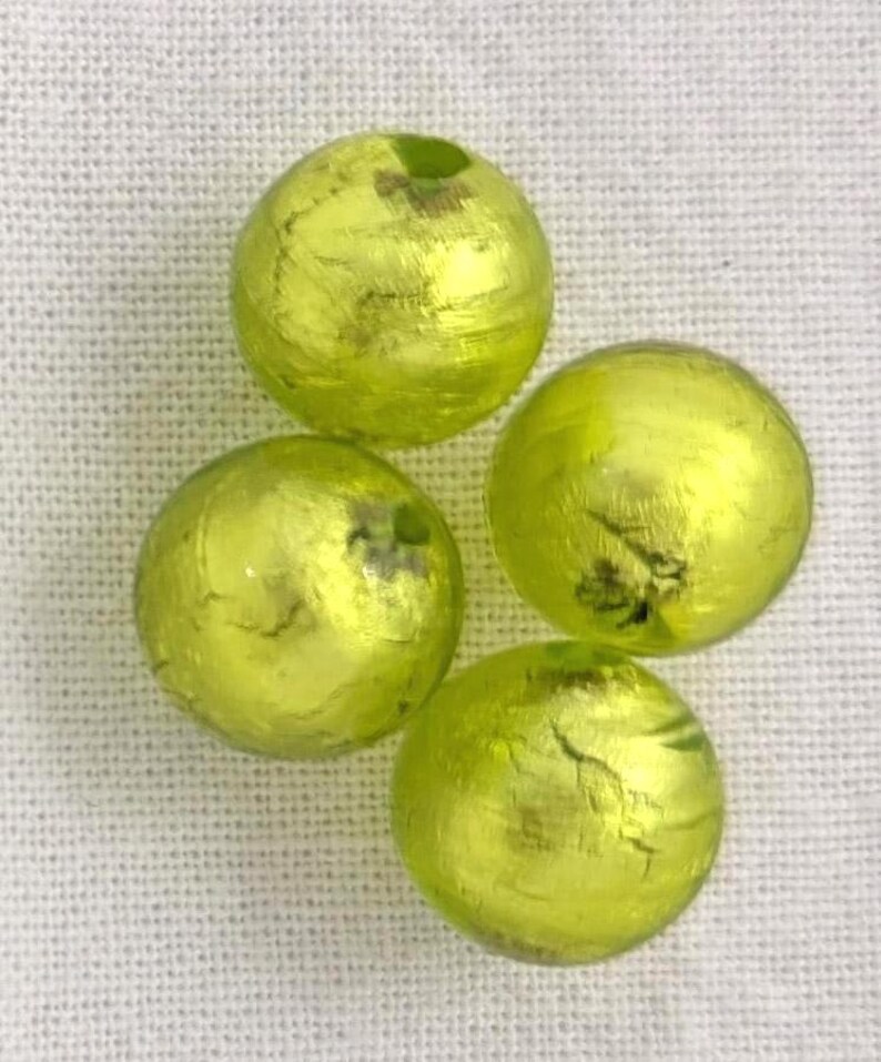 May include: Four round, yellow-green decorative beads with a metallic finish. The beads have a slightly irregular surface and appear to be made of glass or a similar material. They are arranged on a white, textured background.