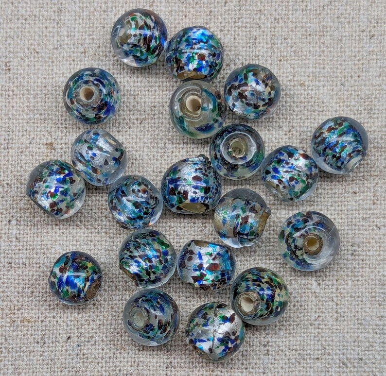 May include: A collection of clear glass beads with a speckled design. The beads feature a mix of blue, green, silver, and brown colors. Each bead has a hole running through the center, suggesting they are for jewelry making or crafts.