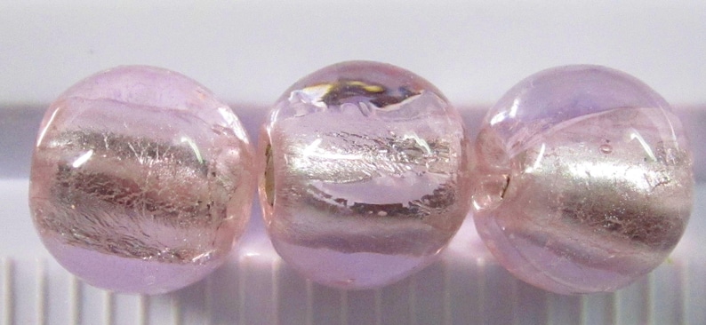 May include: Three round, light pink glass beads with a silver metallic stripe. The beads are translucent, allowing light to pass through. The beads are approximately 0.5 inches in diameter.