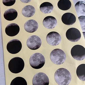 May include: A sheet of stickers featuring different phases of the moon. The stickers are black and silver with a glossy finish.