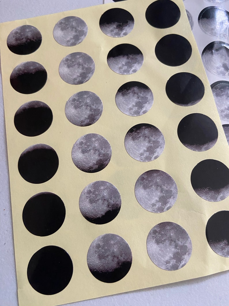 May include: A sheet of stickers featuring different phases of the moon. The stickers are black and silver with a glossy finish.