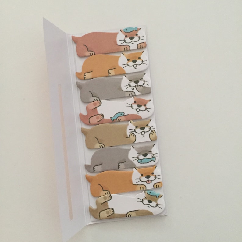 K&ouml;nnte beinhalten: Ein Satz von acht bunten Haftnotizen mit Comic-Ottern mit verschiedenen Gesichtsausdr&uuml;cken. Die Otter halten Fische und tragen H&uuml;te. Die Notizen sind an einer wei&szlig;en Karte befestigt.