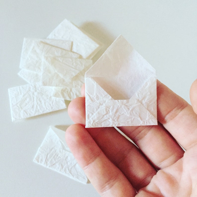 May include: A hand holding a small white envelope with a textured surface. There are several other white envelopes on a white surface.
